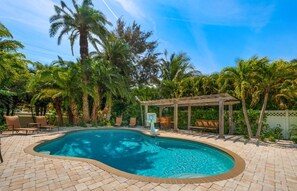A swimming pool with a wooden pergola, surrounded by palm trees and a brick patio.