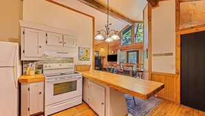 Una cocina con gabinetes blancos, una estufa y una encimera de madera. Hay un comedor con una mesa y sillas, y un letrero que dice "Welcome to Cabin".