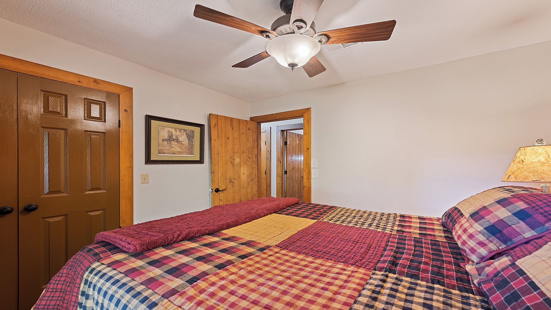 A bedroom with a bed, wooden doors, a ceiling fan, and a framed picture on the wall.