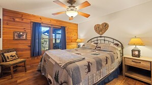 Un dormitorio con pared de paneles de madera, una cama con colcha estampada de animales, un ventilador de techo, una mesita de noche con lámpara y una silla con cojín.