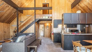 A cozy cabin interior with a living area featuring a sofa and a wooden bench, a kitchen with dark cabinets and stainless steel appliances, and a staircase leading to an upper level. - Chalet #12, Slope Side Ski in Ski Out Spacious Fam (Mammoth Lakes)