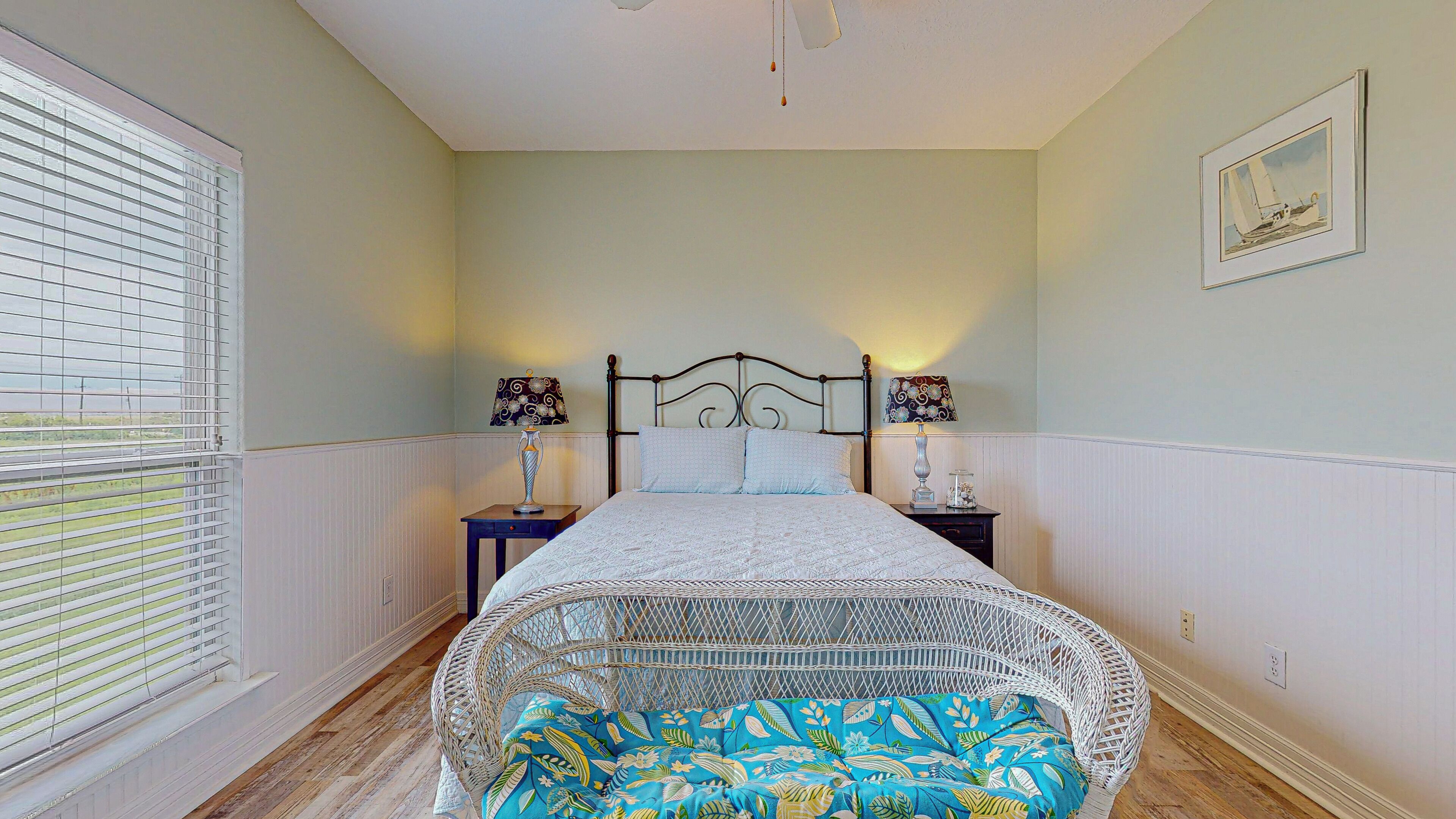A bedroom with a bed, two bedside tables with lamps, a window with blinds, a framed picture on the wall, and a wicker chair with a cushion.