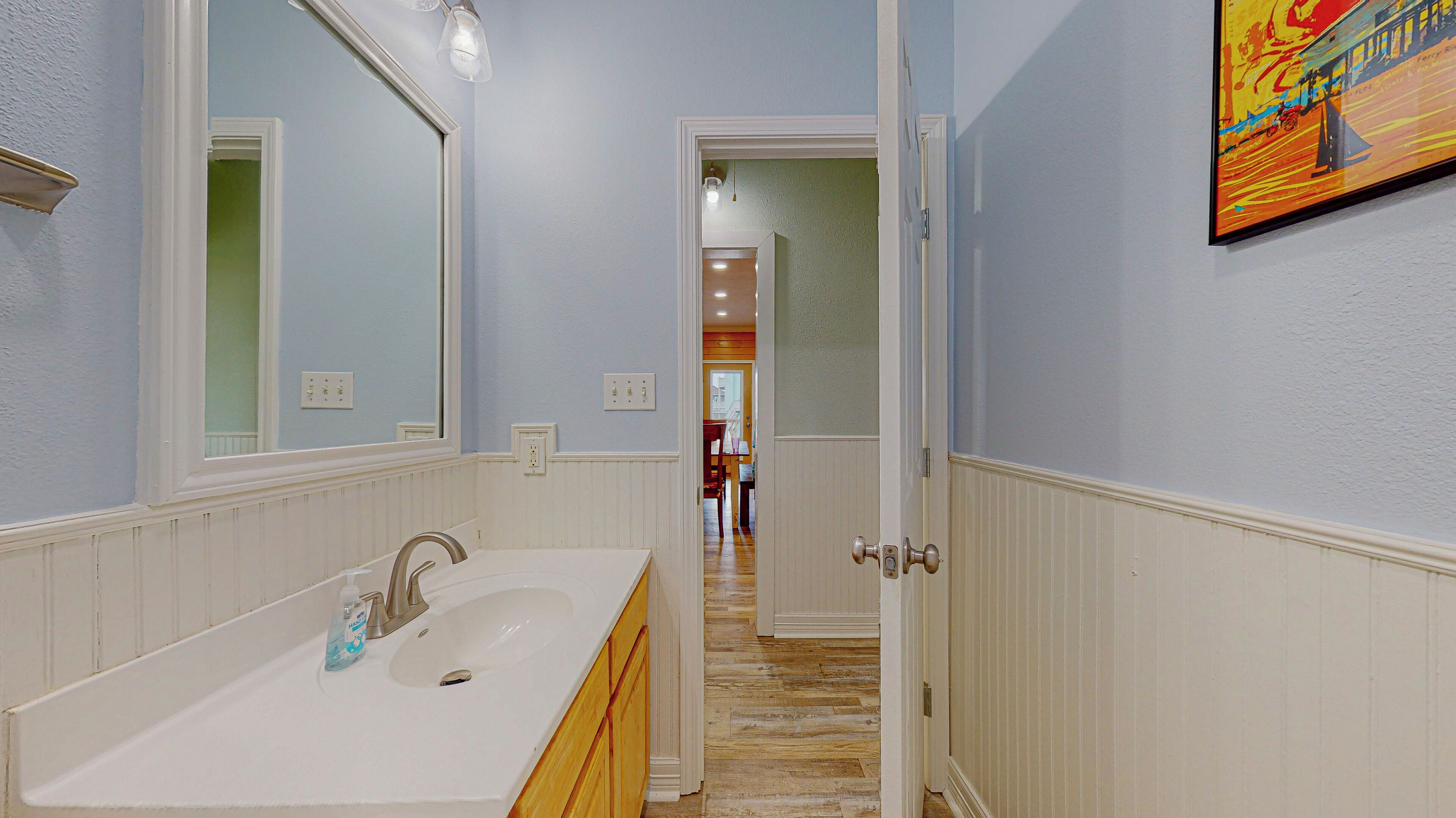 Un couloir avec une salle de bain comprenant un lavabo et un miroir, un plancher en bois et une porte donnant sur une autre pièce.