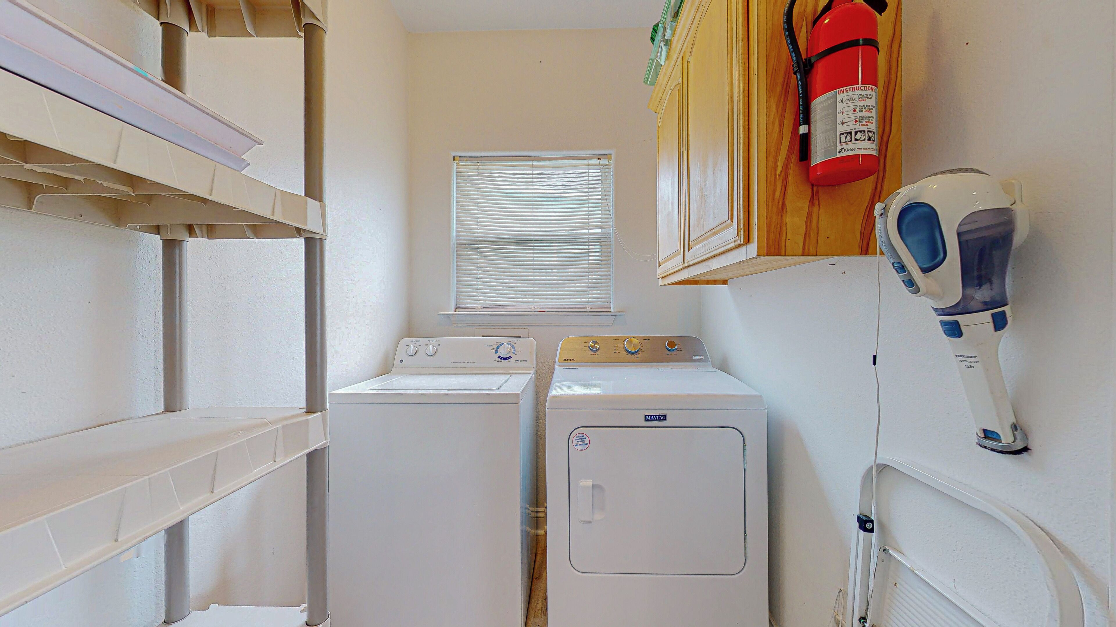 A laundry room with two washing machines, a window with blinds, a fire extinguisher, and a hairdryer.