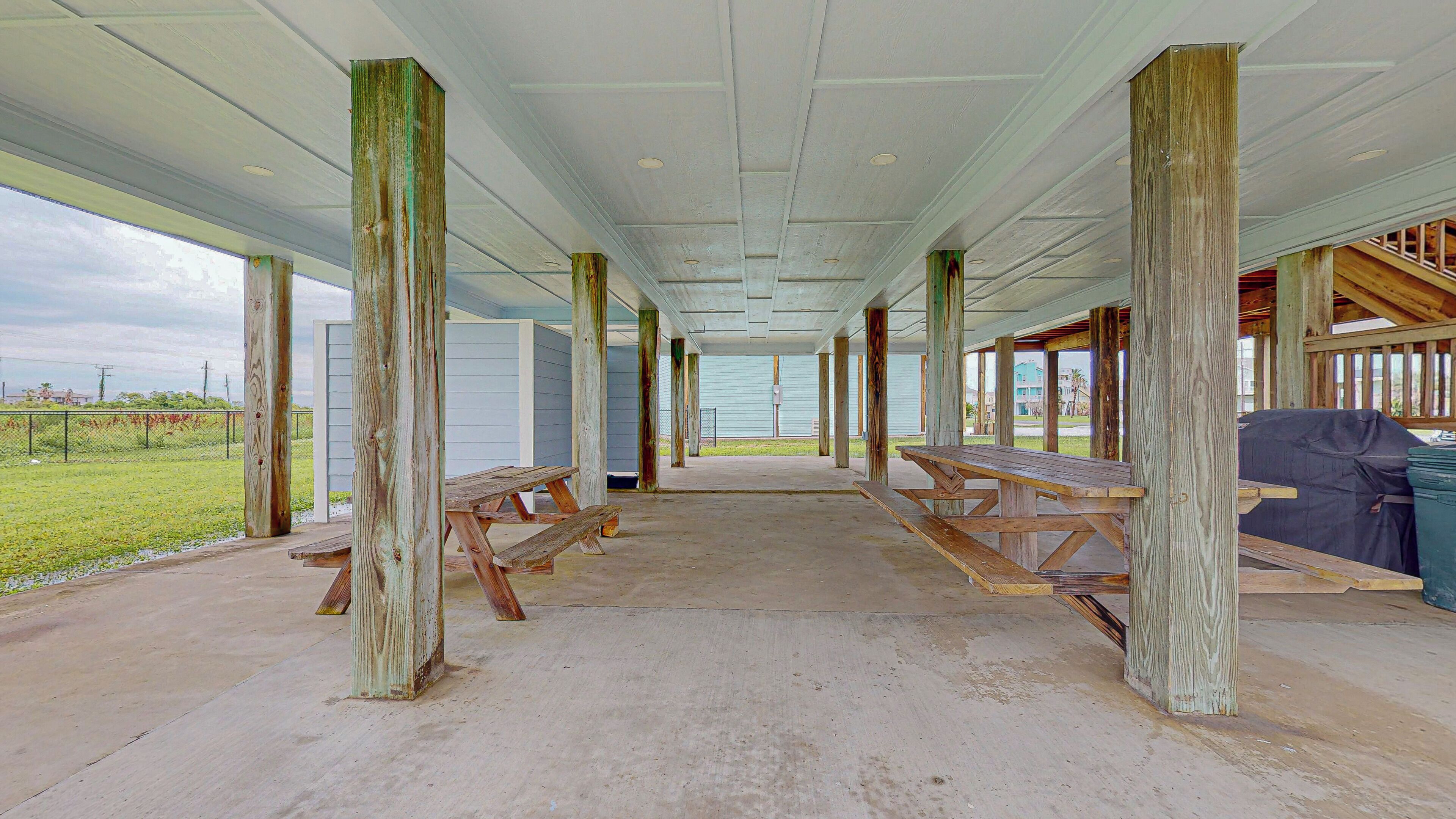An outdoor covered area with picnic tables and wooden posts, a concrete floor, and a view of buildings in the background.