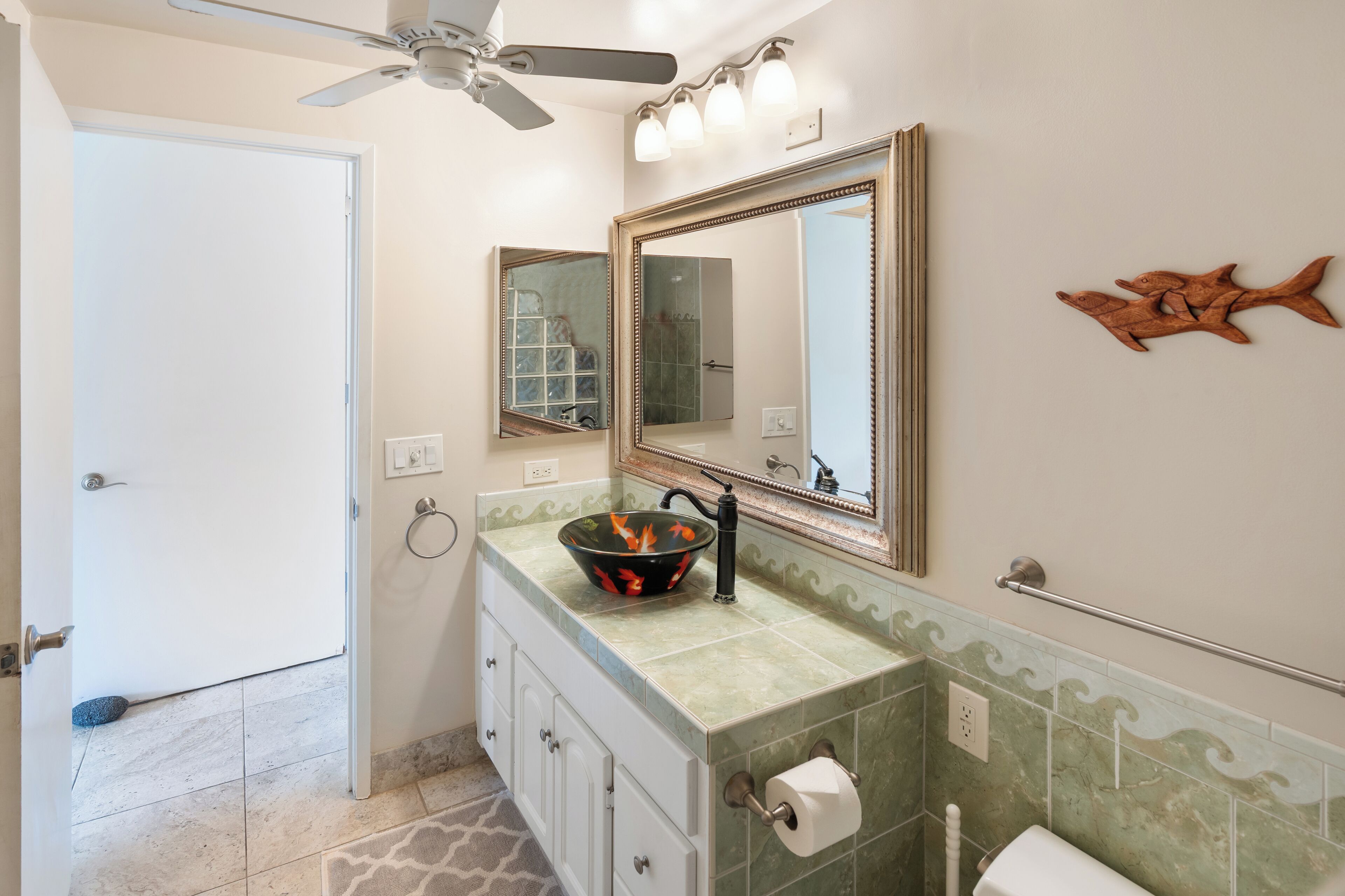 A bathroom with a green tiled vanity, a large mirror, a ceiling fan, and a decorative fish wall art.