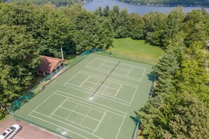 Luftaufnahme eines Tennisplatzes, der von dichten Bäumen umgeben ist, mit einem kleinen Gebäude links.
