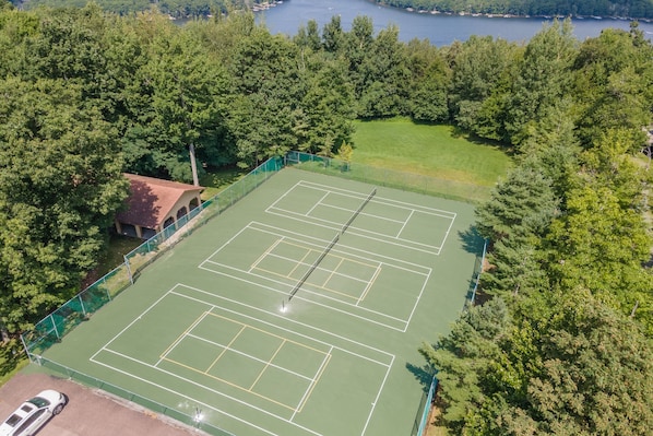 An aerial view of a tennis court surrounded by dense trees, with a small building to the left.