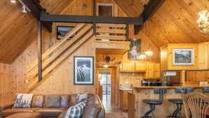 Un intérieur de chalet chaleureux avec un escalier en bois, un coin salon avec un canapé et un pouf, une cuisine avec des tabourets de bar et des tableaux encadrés aux murs.