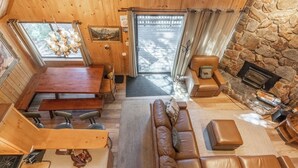 L’intérieur chaleureux d’un chalet avec une cheminée en pierre, un coin repas en bois, un canapé en cuir et une grande fenêtre donnant sur une forêt.
