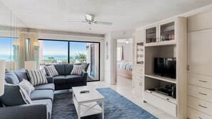A modern living room with a grey sofa, a white coffee table, and a TV on a cabinet. There is a balcony with a view of the ocean. - Beach Palms 203 - 3 Bedroom / 2 Bath - Gulf View Condo (Indian Shores)