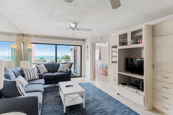 A modern living room with a grey sofa, a white coffee table, and a TV on a cabinet. There is a balcony with a view of the ocean.