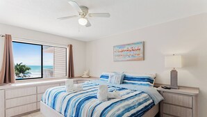 A bedroom with a large bed, a ceiling fan, a painting, a nightstand, and a view of the beach. - Beach Palms 203 - 3 Bedroom / 2 Bath - Gulf View Condo (Indian Shores)