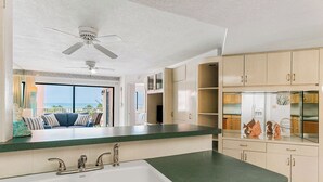 A kitchen with a green countertop, white sink, and beige cabinets. There is a ceiling fan and a view of the ocean through a large window. - Beach Palms 203 - 3 Bedroom / 2 Bath - Gulf View Condo (Indian Shores)