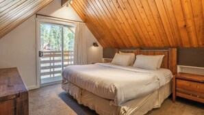 Un dormitorio con techo de madera inclinado, una cama con sábanas blancas, una mesita de noche y vistas a través de un ventanal.