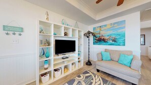 A living room with a TV, white shelving with decorative items, a sofa with teal cushions, and a painting of a seascape. - Aloha Cabana: 2  BR, 1  BA House in Crystal Beach, Sleeps 8 (Crystal Beach)