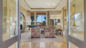 A spacious living room with a large window, a piano, and a sofa set. - FALL GETAWAYS 25% OFF Poolside Perfection 50ft Pool Dreamy Vibes (LA QUINTA)