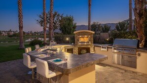 An outdoor kitchen with a stone fireplace, a grill, and bar stools. - FALL GETAWAYS 25% OFF Poolside Perfection 50ft Pool Dreamy Vibes (LA QUINTA)