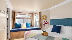 Ein Schlafzimmer mit Bett, einer Sitzbank am Fenster mit Kissen, einem Nachttisch mit Pflanze und einer Wanddekoration.