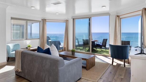 Ein geräumiges Wohnzimmer mit einem grauen Sofa, blauen Sesseln und einem Holztisch. Große Fenster bieten Blick aufs Meer.