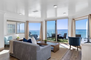 Un salón amplio con un sofá gris, sillas azules y una mesa de centro de madera. Amplias ventanas ofrecen vistas al océano.