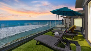 Ein Balkon mit Blick auf das Meer bei Sonnenuntergang, ausgestattet mit Korbstühlen und einem großen Sonnenschirm.
