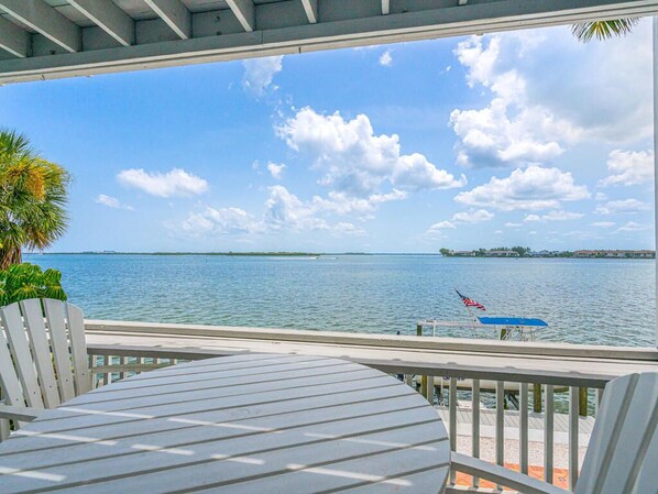 A view from a covered outdoor area with a table and chairs, overlooking a body of water with a distant shoreline and a flag.