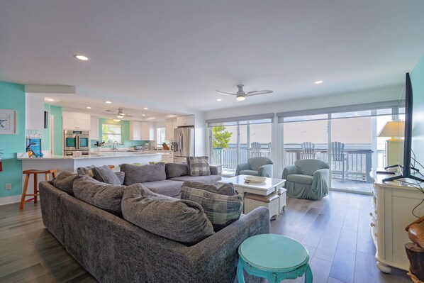 A spacious living area with a grey sectional sofa, a teal coffee table, and a balcony with chairs overlooking a body of water.