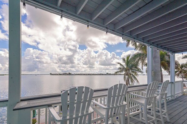 A covered deck with white chairs overlooking a calm body of water and a cloudy sky.