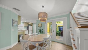 A dining area with a round wooden table and white chairs, a hanging light fixture, and a view of the kitchen and outdoor area through a glass door. - A Time Out: *Quaint Cottage *Heated Pool *Across the street from the Bay! (Anna Maria)