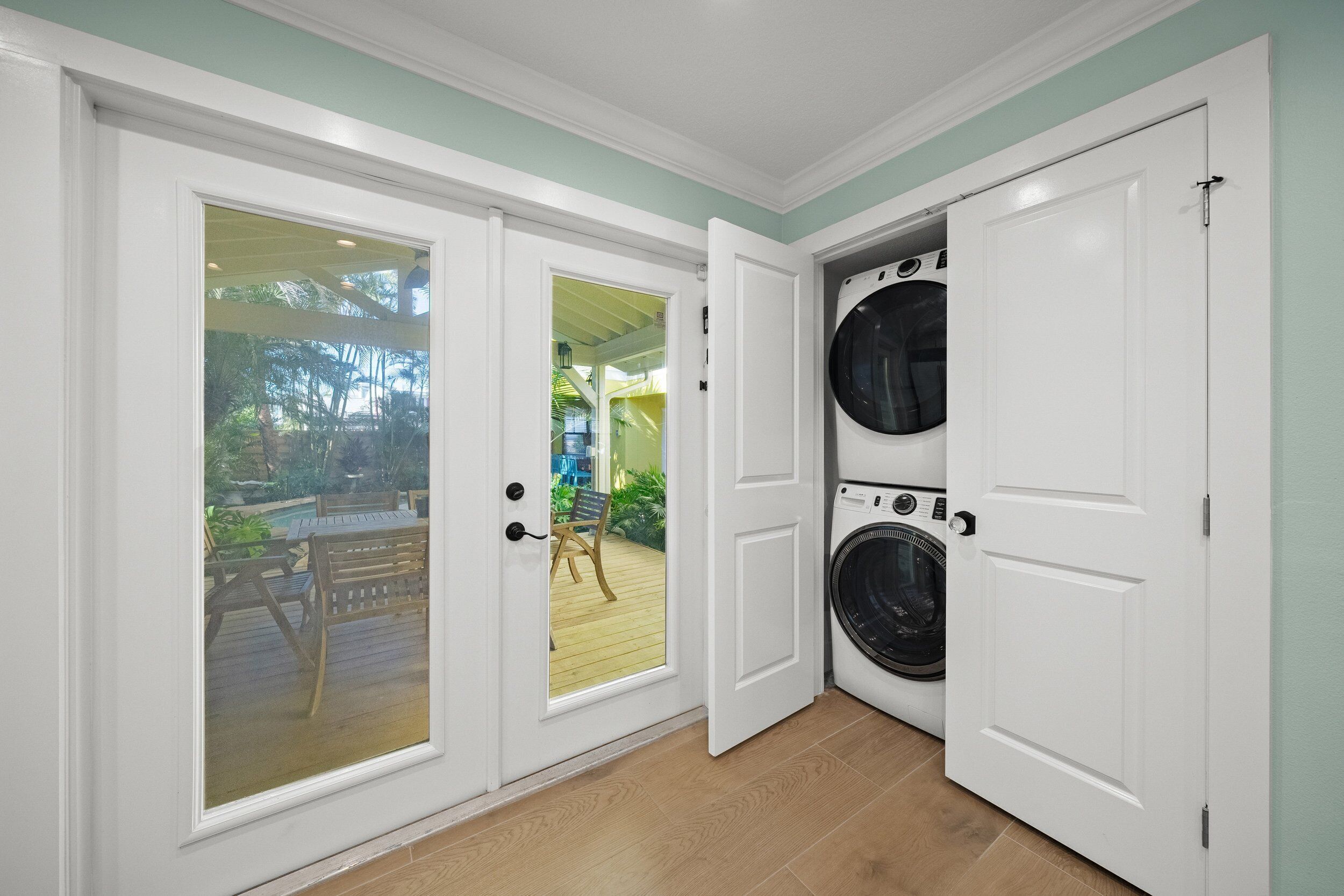 Une buanderie équipée de deux machines à laver, une terrasse en bois visible à travers une porte vitrée et une porte blanche donnant sur un espace extérieur abrité.