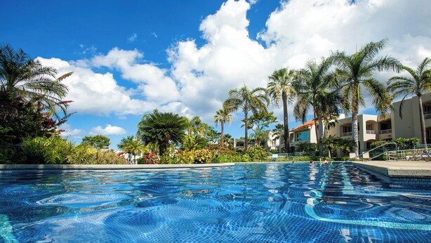 A clear blue pool surrounded by palm trees and a building with balconies.