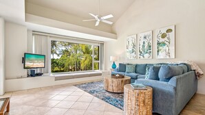 A modern living room with a large window, a ceiling fan, a blue sofa, and a flat-screen TV.