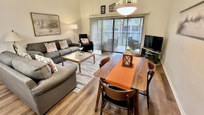 Ein Wohnzimmer mit grauem Sofa, einem Holzesstisch und einem Fernseher auf einem Schrank.
