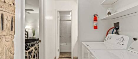 A laundry room with a Whirlpool washer and dryer, a fire extinguisher on the wall, and a doorway leading to a bathroom.