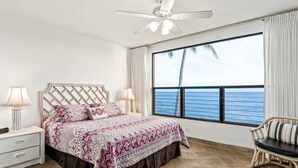 Une chambre à coucher comprenant un lit, des tables de chevet, une chaise, un ventilateur de plafond et une grande fenêtre donnant sur l’océan.