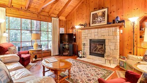 L’intérieur chaleureux d’un chalet en bois, avec une cheminée en pierre, des murs en bois et une grande fenêtre donnant sur une forêt.
