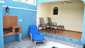 Un área de piscina con una silla de descanso azul, una parrilla y una zona de estar con sillas y una mesa.