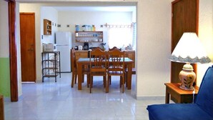 Un comedor con una mesa y sillas de madera, una cocina al fondo y una lámpara sobre una mesita.