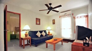 Una sala de estar con un sofá azul, dos mesas auxiliares con lámparas, una mesa de centro de madera, un ventilador de techo, un televisor y una puerta que da a otra habitación.