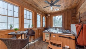 A wooden cabin interior with a hot tub, wicker chairs, and large windows.