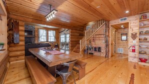 A log cabin interior with a wooden dining table, benches, a fireplace, a staircase, and a shelf with various items.