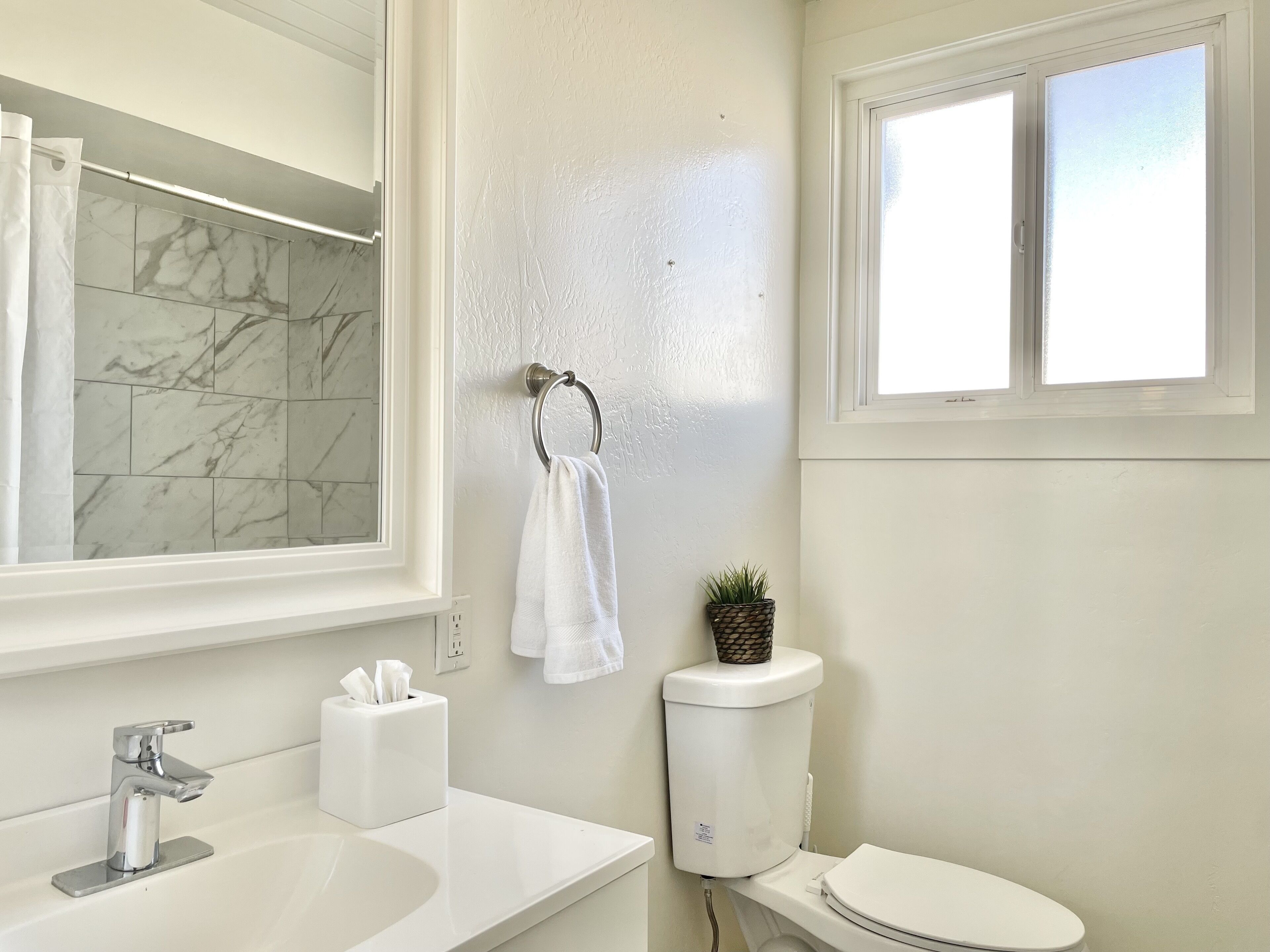 Un baño con ducha de azulejos de mármol, un lavamanos blanco, inodoro y ventana.