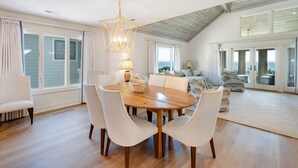 A spacious dining area with a wooden table and white chairs, a chandelier, and large windows offering a view of the outdoors.