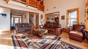 Ein großzügiges Wohnzimmer mit hoher Decke, Holzdielen und einem großen Teppich mit Tiermotiven. Mehrere Ledersofas und ein Couchtisch aus Holz sind vorhanden.