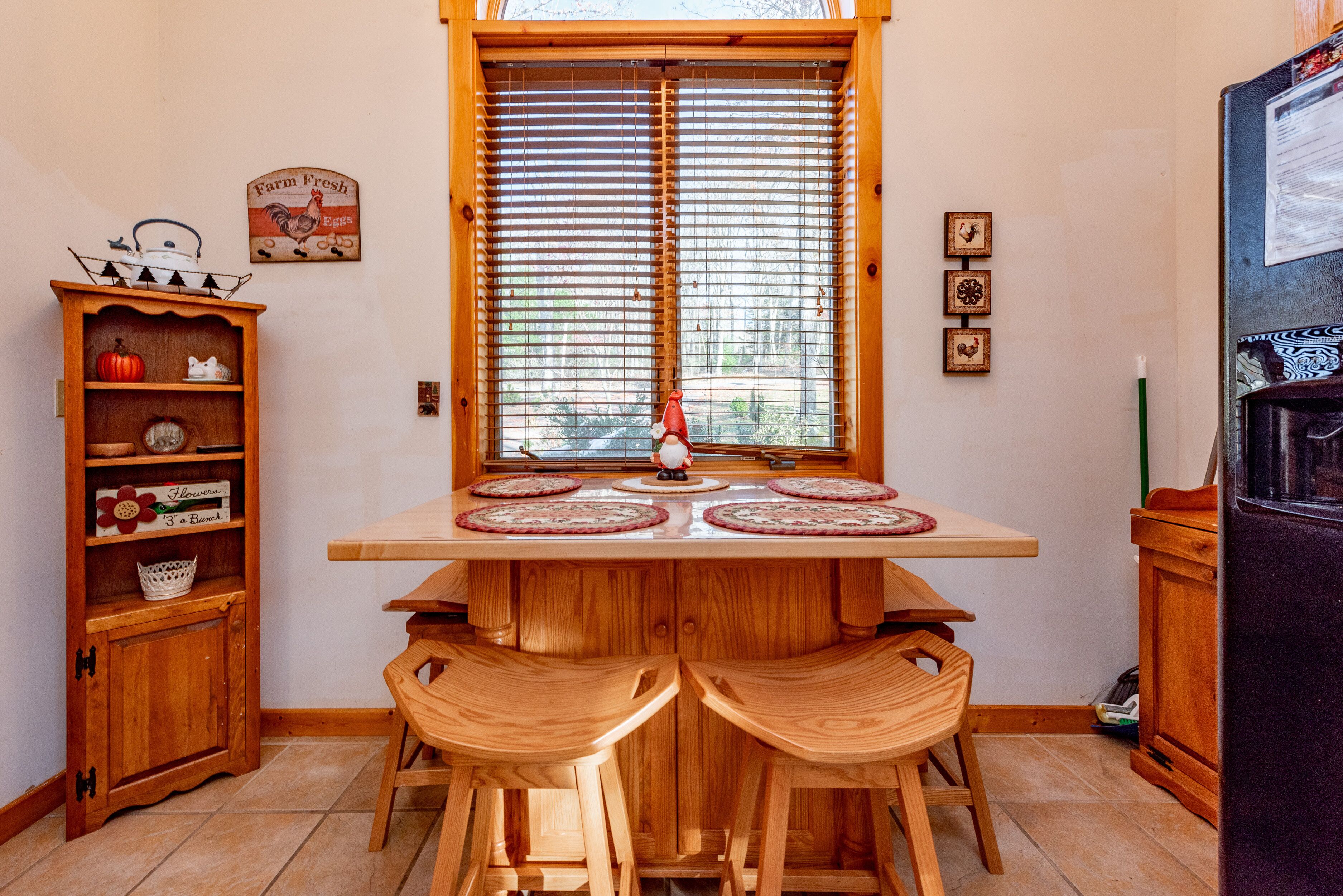 A kitchen with a wooden table and two stools, a window with blinds, and a refrigerator.
