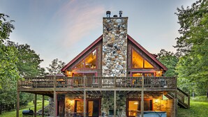 A two-story log cabin with a stone chimney, wooden deck, and outdoor seating area. - Dog Friendly Home in Deep Creek Hills w/ Hot Tub (McHenry)