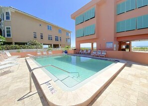 A swimming pool with lounge chairs, a building with balconies, and a clear sky.