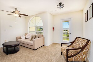 A living room with a beige sofa, a wooden coffee table, a patterned armchair, a ceiling fan, and a door leading outside.