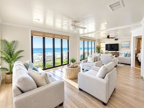 A spacious living room with a large window overlooking the beach, a ceiling fan, and a flat-screen TV. - Sand, Sun, Serenity P1111-23 (Oceanside)